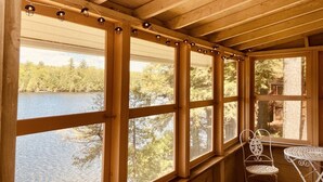 Una cabaña con ventanas grandes que dan a un lago, una silla metálica y una mesita.