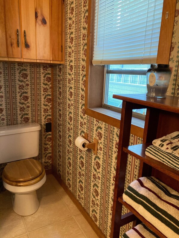 Un baño con inodoro, armarios de madera, una ventana con persianas, un dispensador de papel higiénico y un estante con una toalla.
