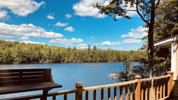 Una terraza de madera con banca y mesa da a un lago, rodeado de árboles y un cielo despejado.