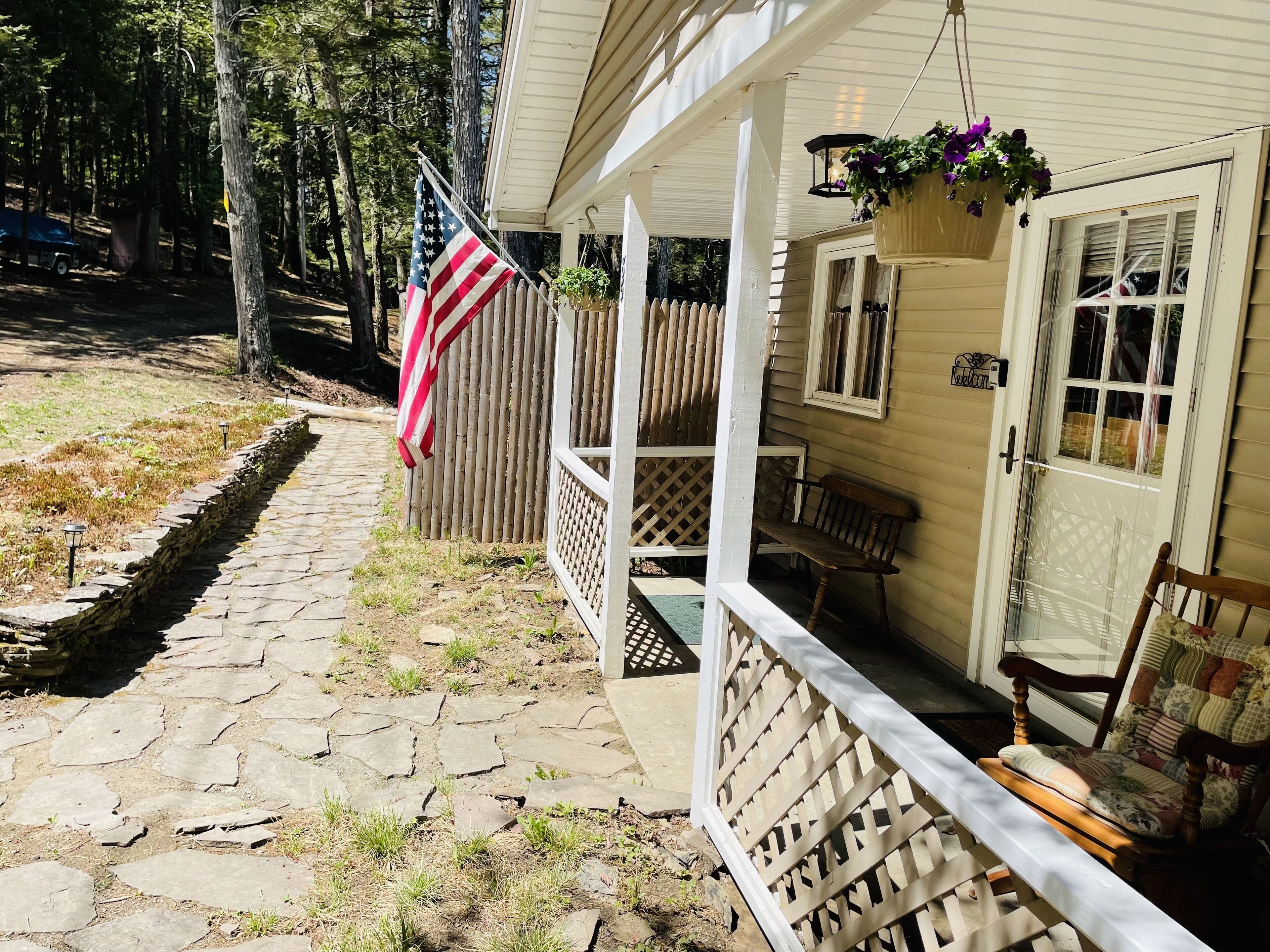 A covered porch with a wooden bench, a hanging flower pot, and an American flag.