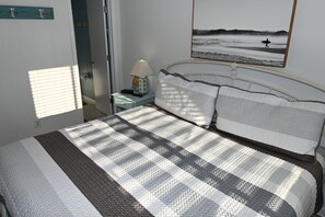 Un lit bien fait, recouvert d’une couvre-lit à rayures, une table de chevet avec une lampe et un tableau encadré au mur.