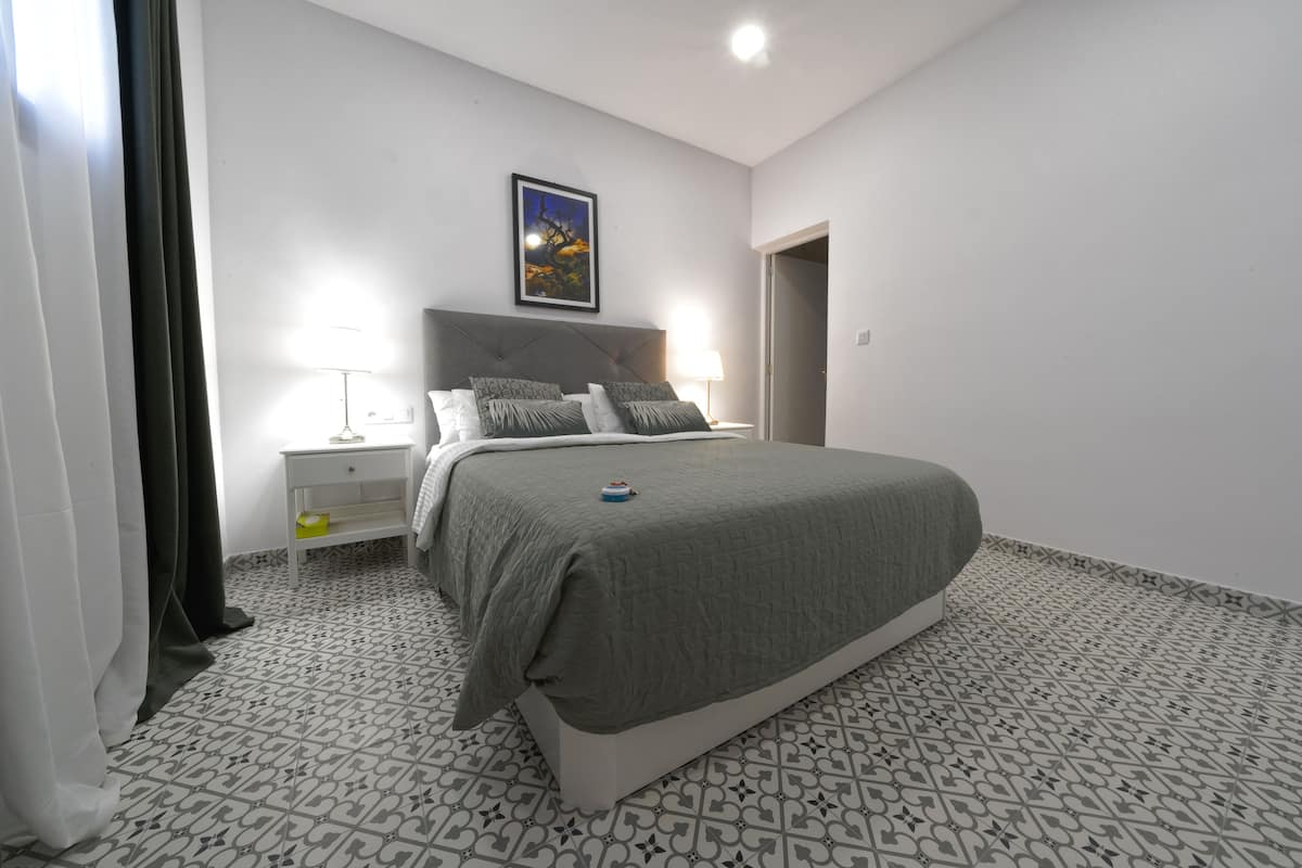 A bedroom with a bed, nightstands, a lamp, a picture on the wall, and patterned floor tiles.