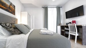 A neatly made bed with a grey bedspread, a white pillow, and a rolled-up towel. There is a white chair and a desk with a red coffee machine. A flat-screen TV is mounted on the wall. - CASA VERDE Comfortable Air-Conditioned (Ingenio)