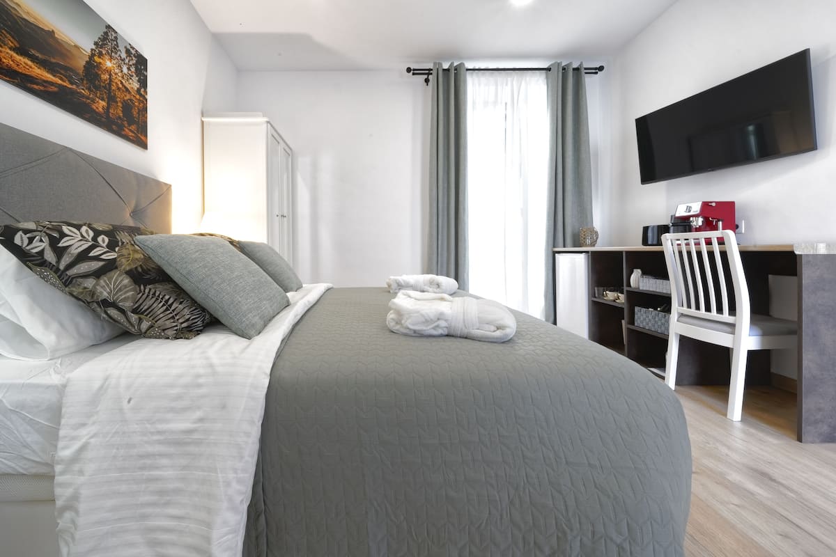 A neatly made bed with a grey bedspread, a white pillow, and a rolled-up towel. There is a white chair and a desk with a red coffee machine. A flat-screen TV is mounted on the wall.