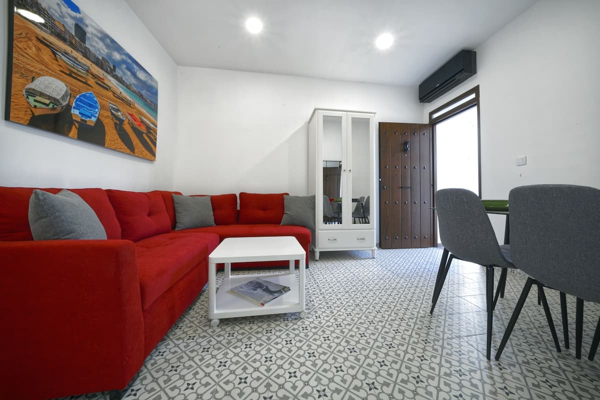A living room with a red sofa, a white coffee table, and a grey armchair. There is a cabinet with glass doors and a painting of boats on the wall.