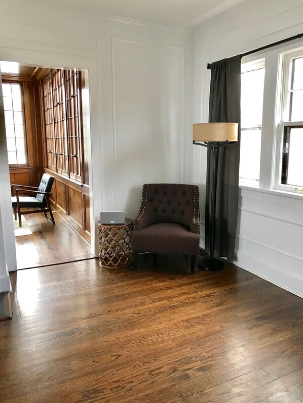Ein Zimmer mit Holzboden, einem braunen Sessel, einem Beistelltisch und einer Lampe. Ein Fenster mit Vorhängen bietet Blick auf eine Holzkonstruktion draußen.