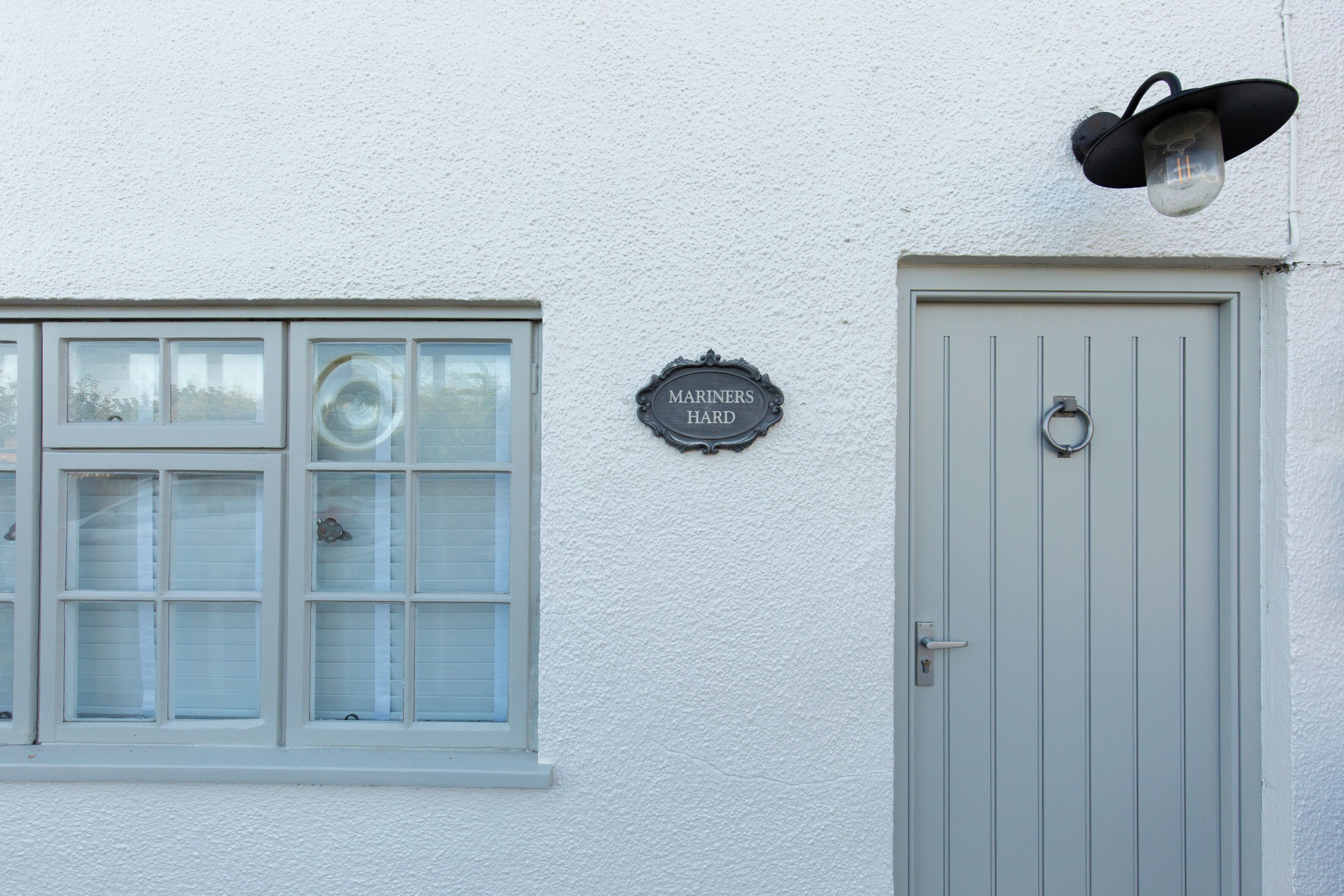 A building with a white exterior, a window with blinds, a door with a handle, and a sign reading 'MARINERS HARD'.