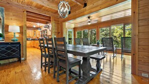 A wooden cabin interior with a dining area, kitchen, and large windows.