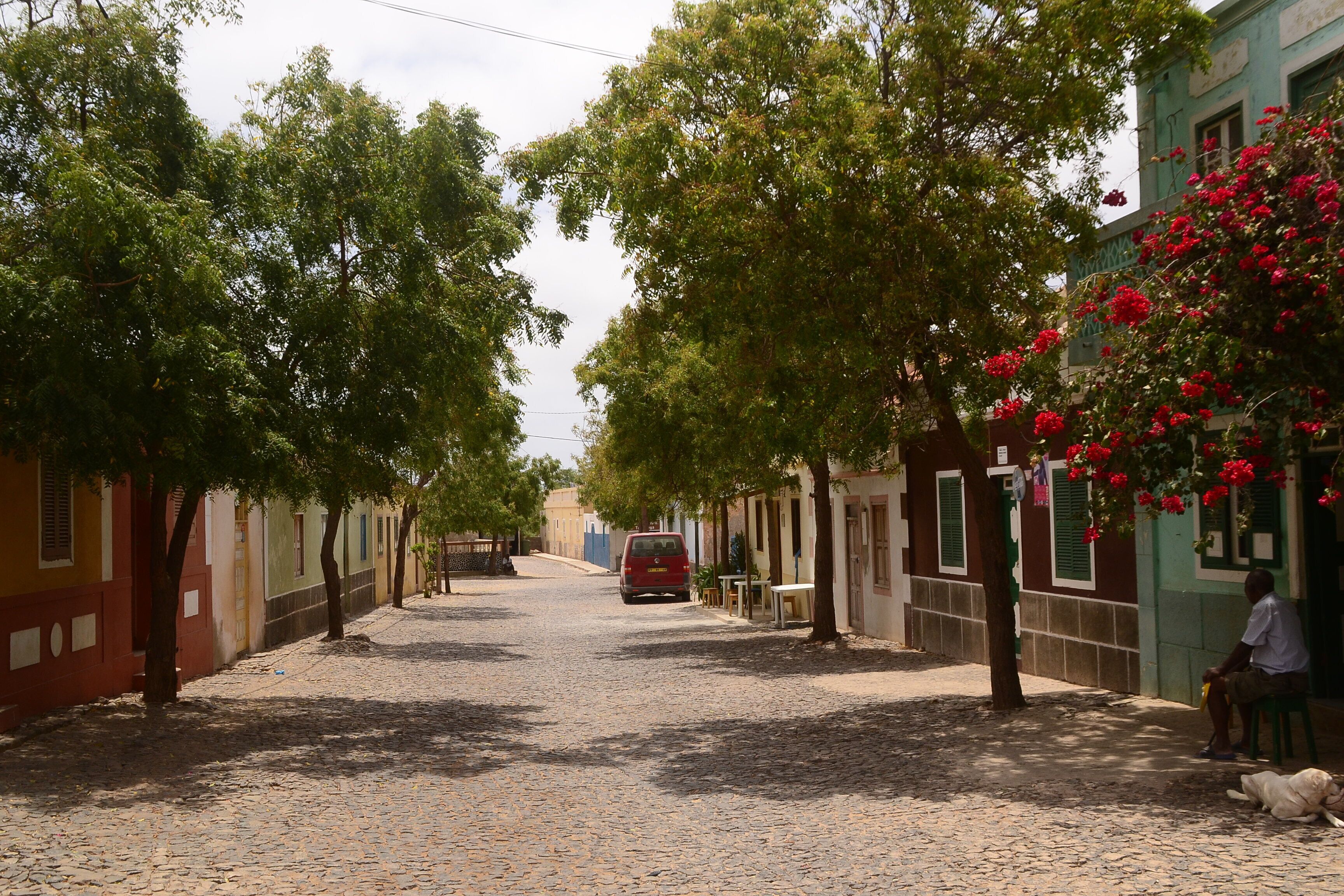 A cobblestone street lined with colorful buildings, a red car parked on the road, and a person sitting on a bench with a dog.