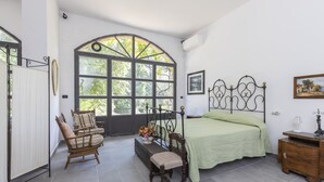 A bedroom with a large arched window, a bed with a green bedspread, a wooden bench, a small table with a vase of flowers, and a wooden nightstand. - Eagle House (CHIANCIANO)