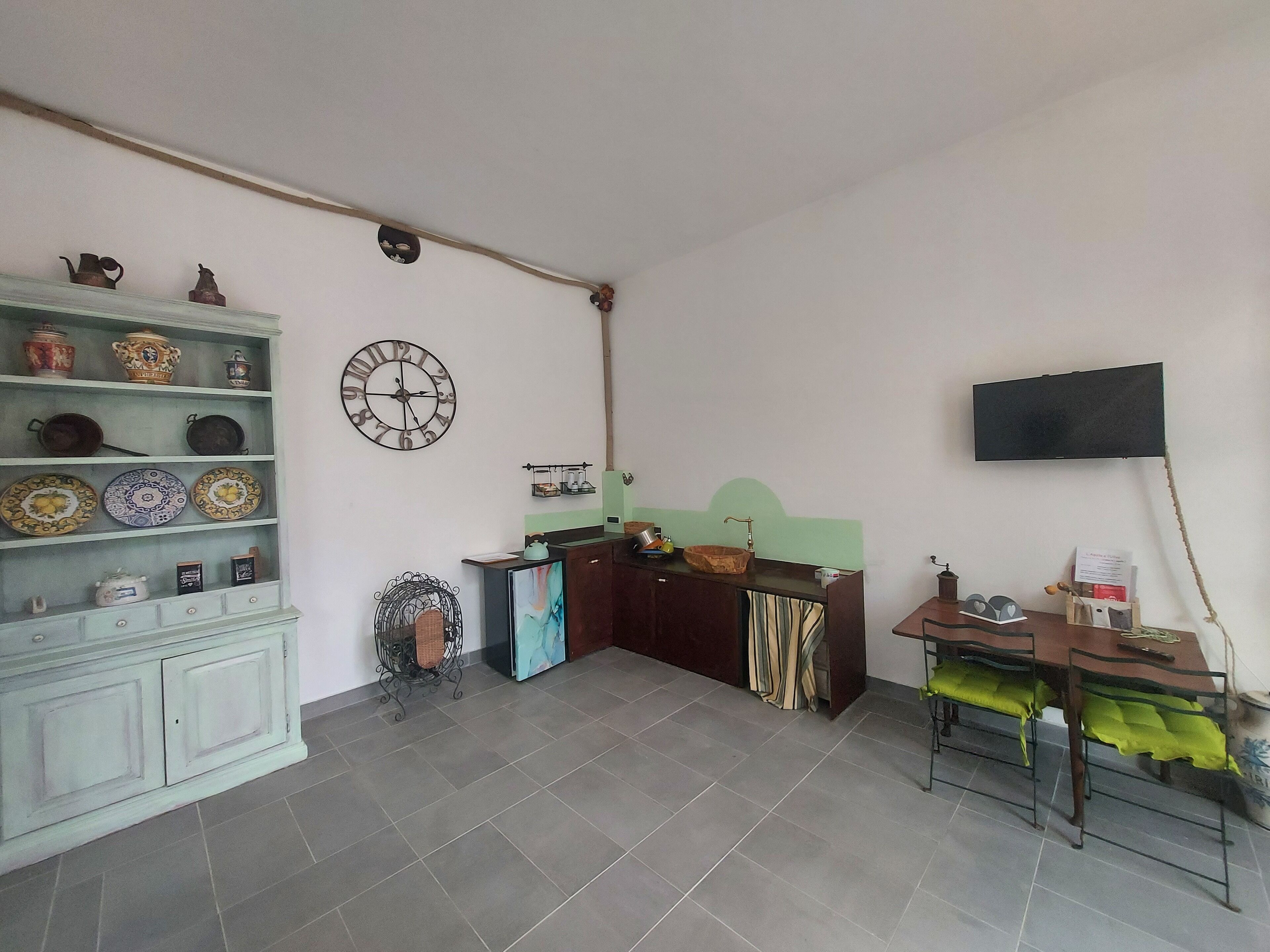 Una habitación con un armario blanco con vajilla, un reloj de pared, un televisor y una pequeña mesa con una silla.