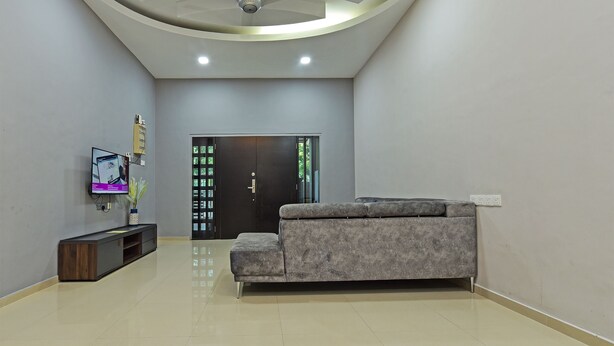 A modern living room with a grey sofa, a flat-screen TV mounted on the wall, a ceiling fan, recessed lighting, and a dark wooden door with glass panels.
