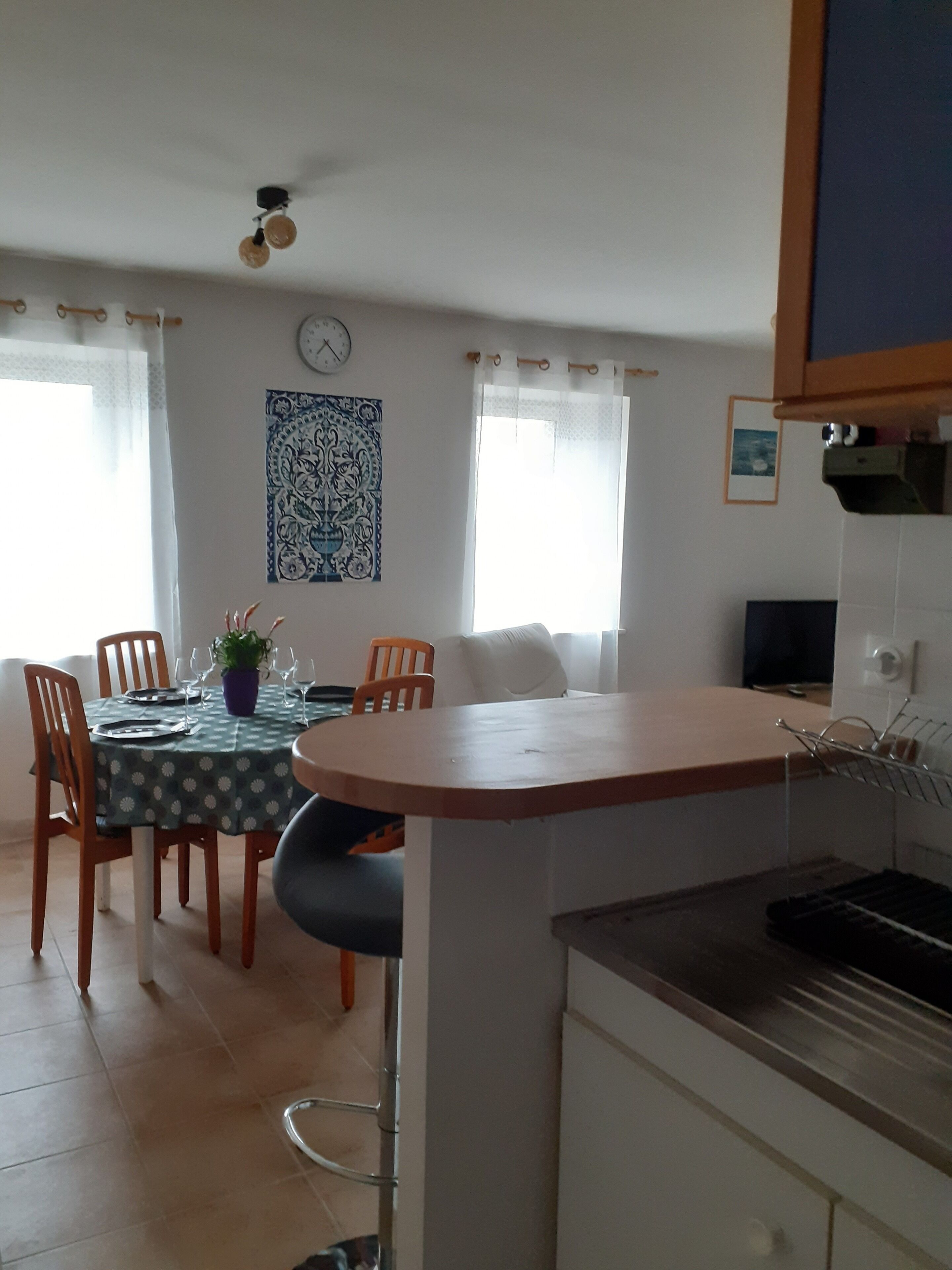 Een eethoek met een tafel voor vier personen, een potplant en een klok aan de muur.
