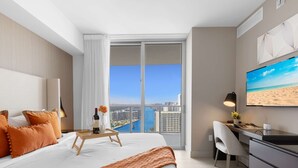 Quarto de hotel com uma janela grande, uma cama com almofadas laranja, uma escrivaninha com cadeira e uma TV exibindo uma cena de praia.