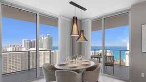 Uma área de refeições moderna com uma mesa redonda posta para quatro pessoas, grandes janelas com vista para a cidade e uma luminária pendente no teto.