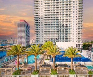 Un hôtel de grande hauteur doté de nombreux balcons, d’un espace piscine agrémenté de palmiers et offrant une vue sur un ciel au coucher du soleil.