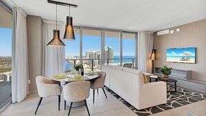 Un salon moderne comprenant un coin repas, de grandes fenêtres offrant une vue sur la ville, un téléviseur à écran plat et un balcon donnant sur le paysage urbain.