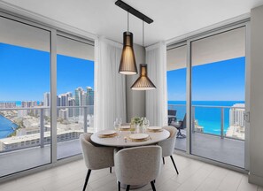 A dining area with a round table, four chairs, and a view of the ocean and cityscape through large windows.