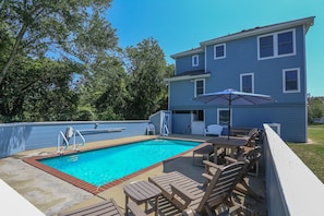 A rectangular swimming pool with a brick edge, surrounded by a concrete deck. There are wooden lounge chairs and a table with a blue umbrella. A two-story building with blue siding and white trim is adjacent to the pool. - The Porter House - Oceanside 6 BR Home in the Heart of Duck - Private Pool, Dog Friendly (Duck)