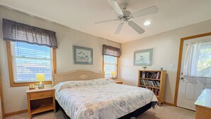 Ein Schlafzimmer mit einem Bett, Nachttischen, einem Bücherregal, einem Deckenventilator und zwei mit Vorhängen versehenen Fenstern.