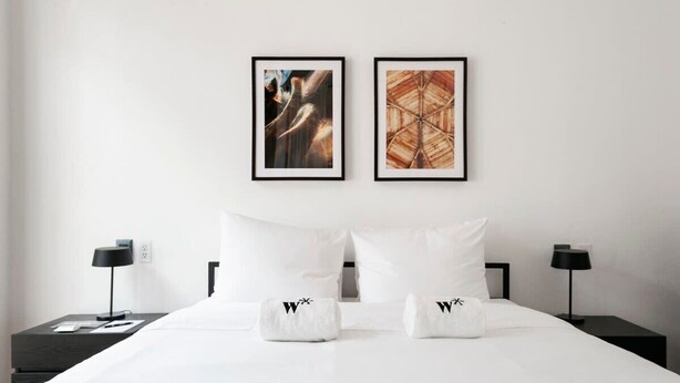 Een hotelkamer met een groot bed, twee nachtkastjes, twee ingelijste kunstwerken en witte handdoeken met een logo.