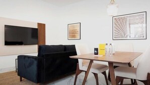 Een moderne woonkamer met een zwarte bank, een houten eettafel met witte stoelen, een wandtelevisie en omlijste kunstwerken.