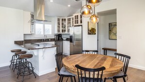 A modern kitchen with white cabinetry, a central island with stools, a round wooden dining table, and pendant lighting. - Luxury Living at the Beach (Stinson Beach)