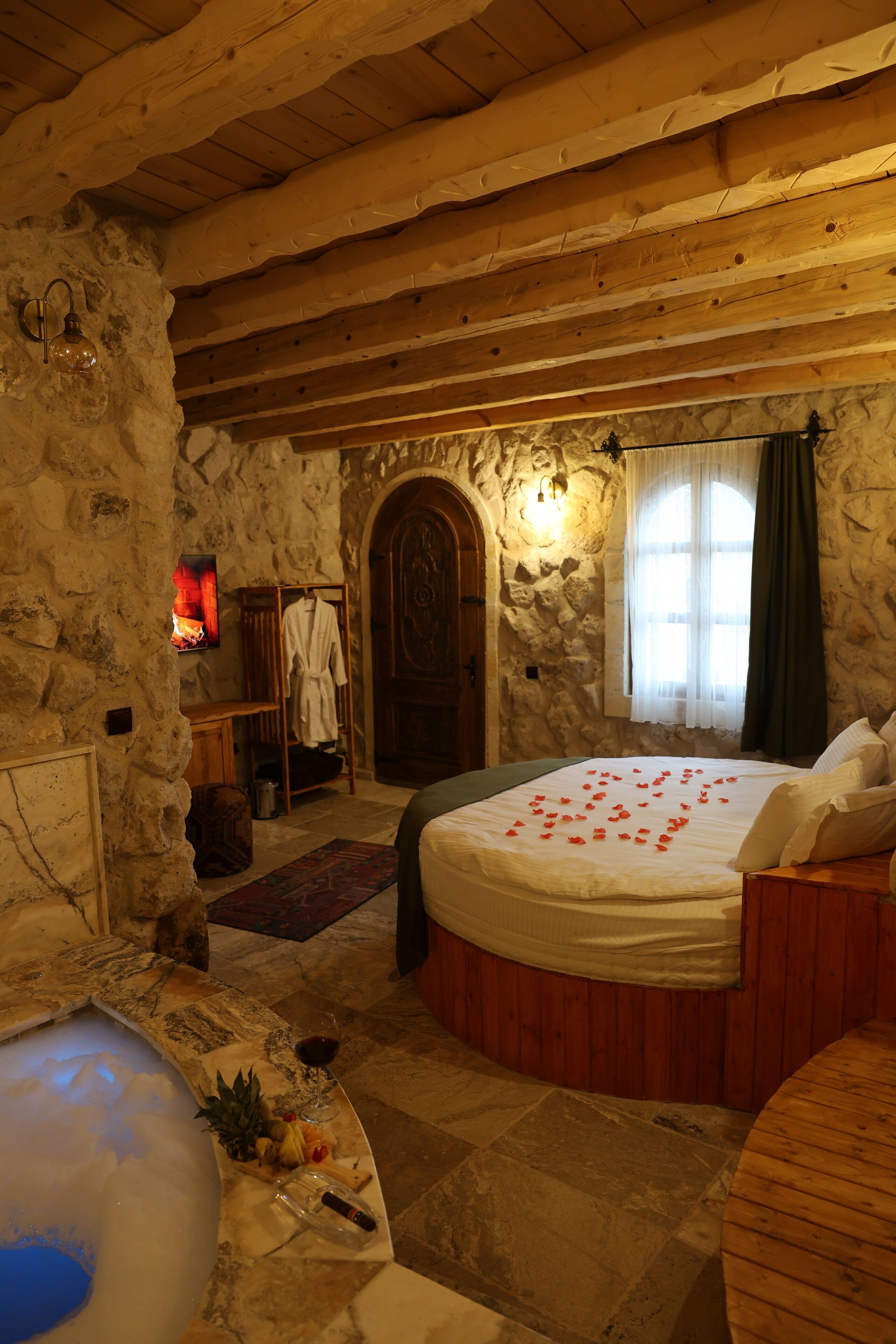 A stone-walled bedroom with a wooden ceiling, a bed with rose petals, a bathtub, and a wooden bench.