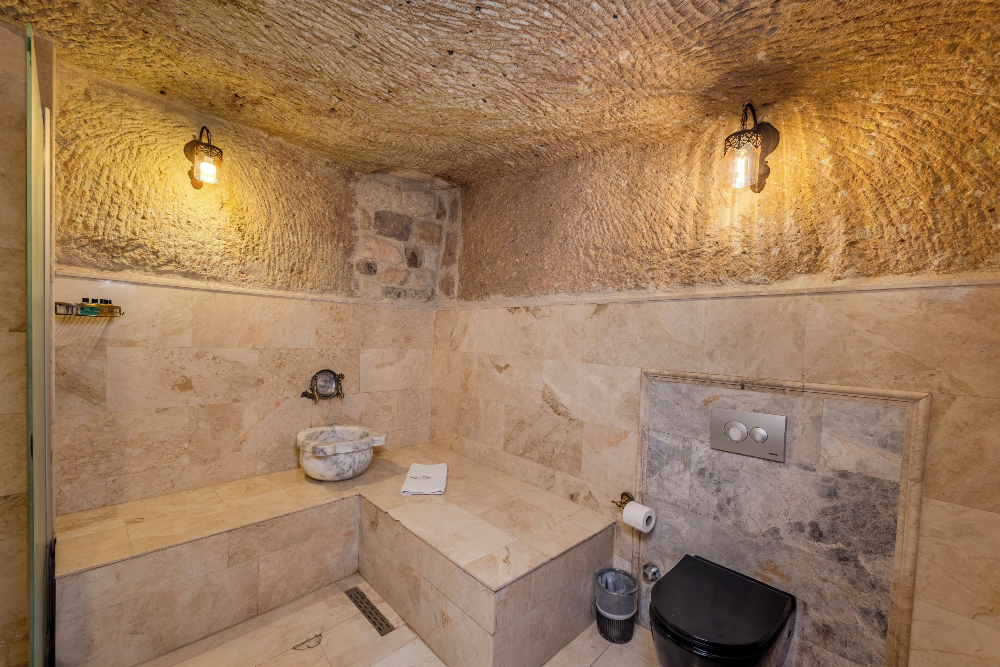 Un baño con paredes de piedra, un lavamanos de mármol, un cubo de basura negro y una toalla sobre el mostrador.