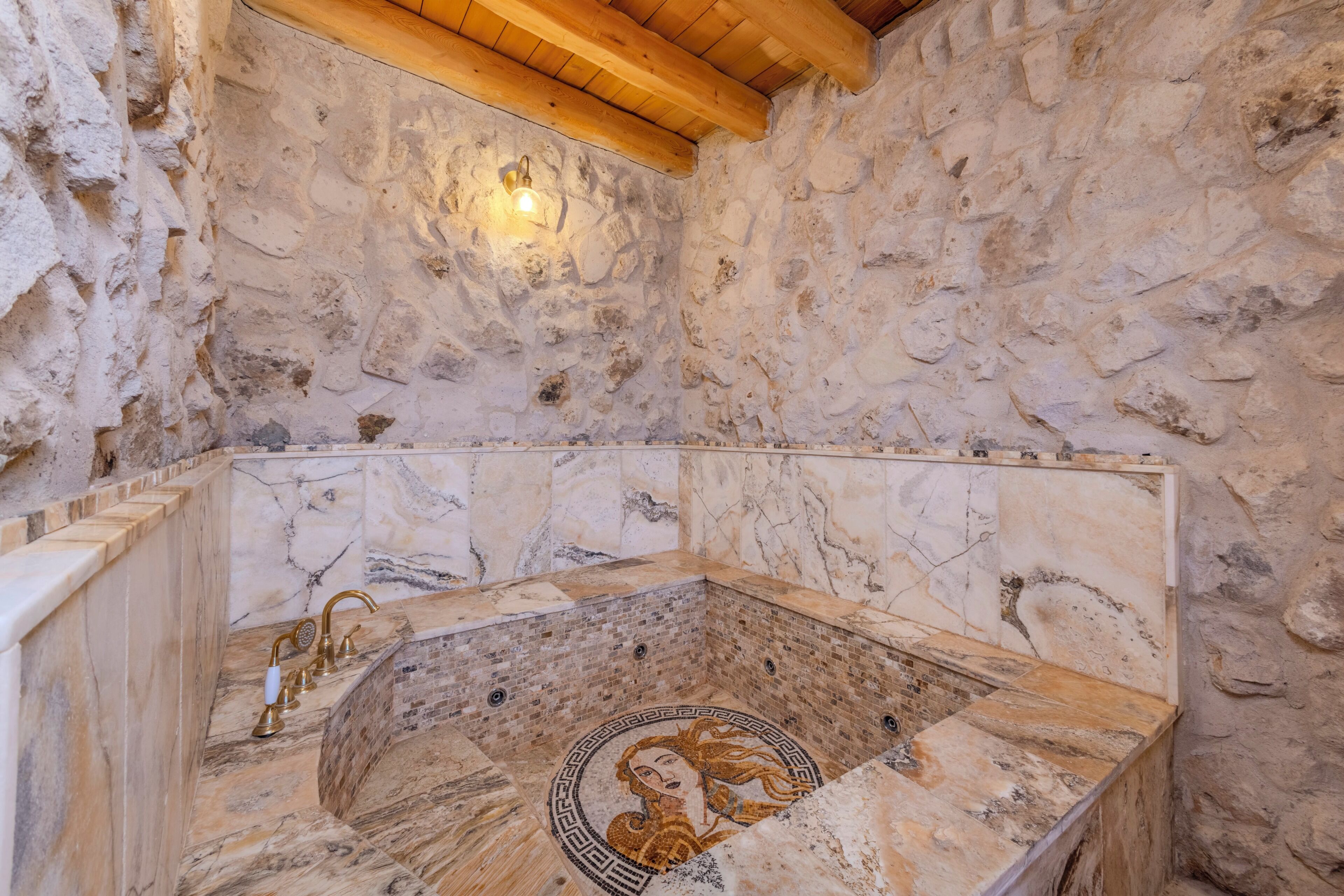 Un baño con paredes de piedra, una bañera de mármol, techo de madera y una lámpara fijada a la pared.