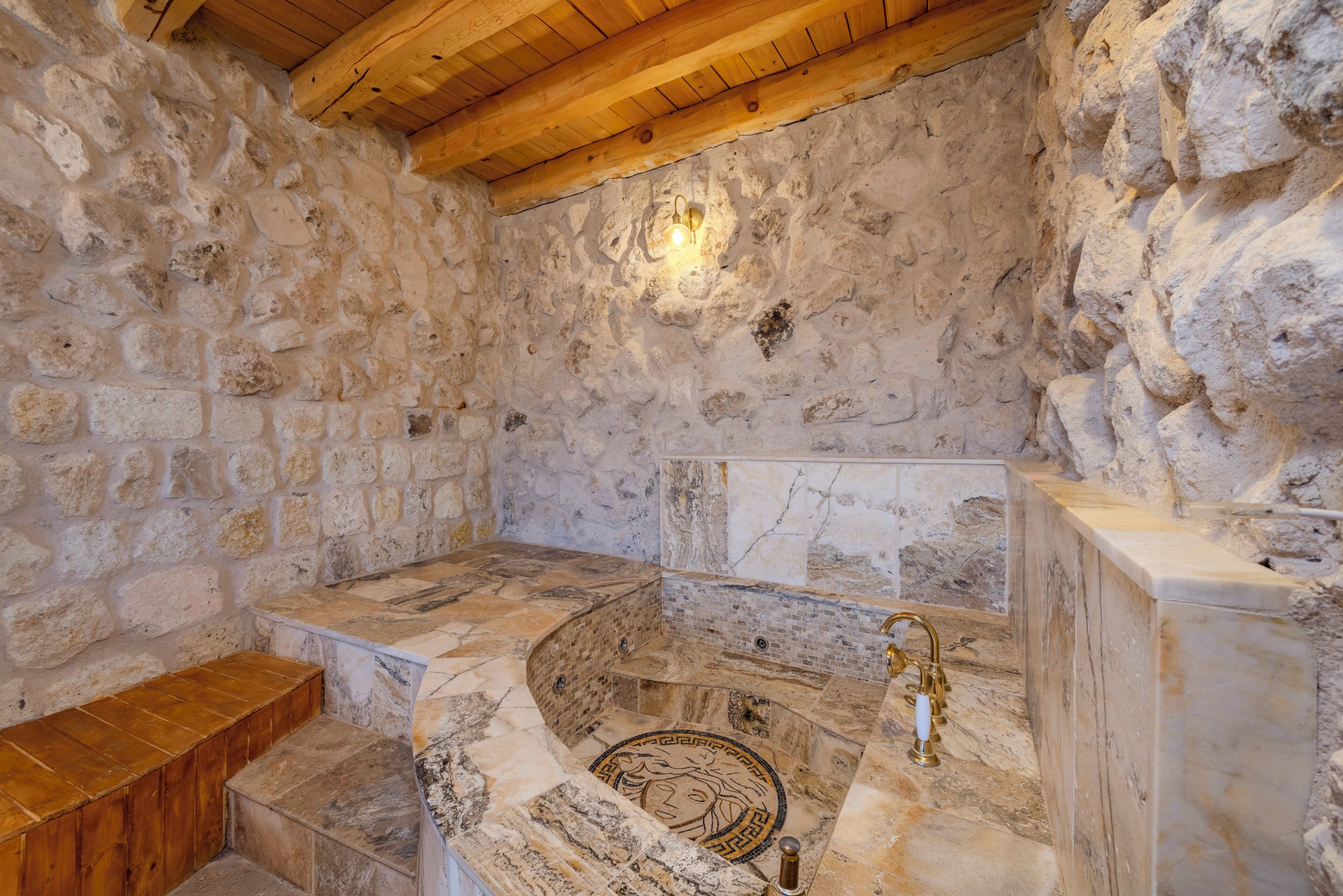 A stone-walled room with a marble bathtub, wooden steps, and a light fixture.