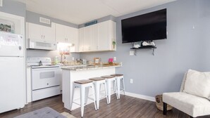 A modern kitchen with white cabinets, a built-in oven, a microwave, and a granite countertop. There are three bar stools, a gray ottoman, and a flat-screen TV mounted on the wall. - One Bedroom, One Bathroom Condo on the Oceanside (Carolina Beach)