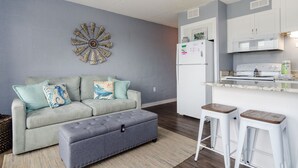A modern living room with a grey sofa, ottoman, and a decorative wall piece. Adjacent is a kitchen with white cabinets, a refrigerator, and a stove. - One Bedroom, One Bathroom Condo on the Oceanside (Carolina Beach)