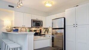 Una cocina moderna con armarios blancos, un refrigerador de acero inoxidable, un microondas y un lavamanos. Hay dos taburetes blancos y un bote de basura.