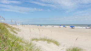 Una playa con dunas de arena, una torre de salvavidas y gente relajándose bajo sombrillas.