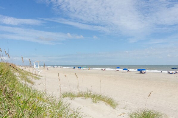 A beach with sand dunes, a lifeguard tower, and people relaxing under umbrellas.