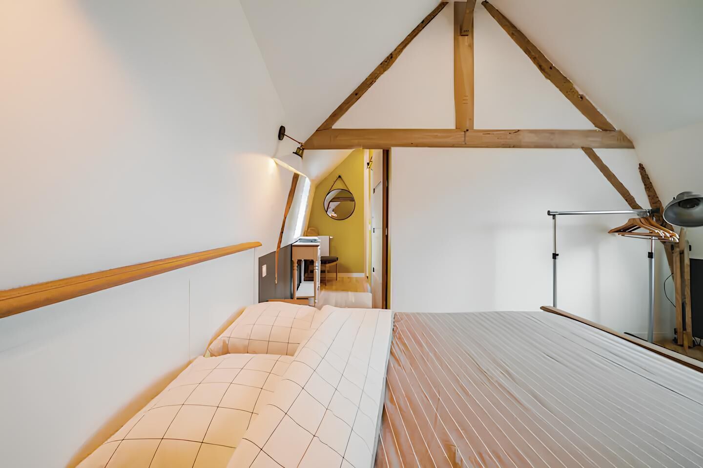 A bedroom with a sloped ceiling, wooden beams, a bed with a striped mattress, a bedside table, and a hanging light fixture.