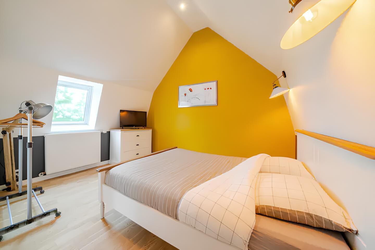 Un dormitorio con techo inclinado, cama, cómoda, televisor y ventana.