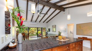 A modern kitchen with granite countertops, wooden cabinets, and a large island. There is a ceiling fan, pendant lights, and a view of the outdoor patio.