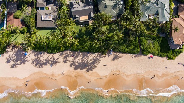 An aerial view of a beachfront property with a sandy beach, clear turquoise waters, and a cluster of palm trees.