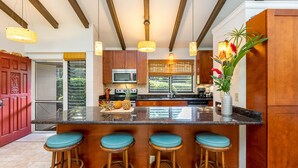 A kitchen with wooden cabinets, a central island with stools, a microwave, and a window with blinds.