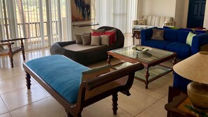 Een ruime woonkamer met een bank, een blauwe zithoek, een glazen salontafel en een houten stoel.