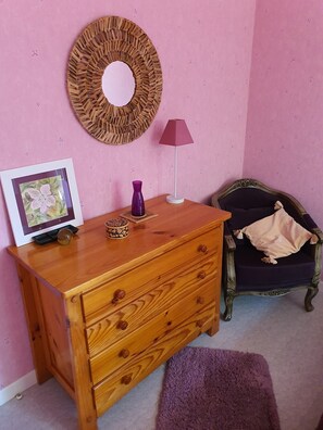 A room with a wooden dresser, a purple vase, a framed picture, a round wall decoration, a floor lamp, a dark blue chair with a beige throw, and a purple rug.