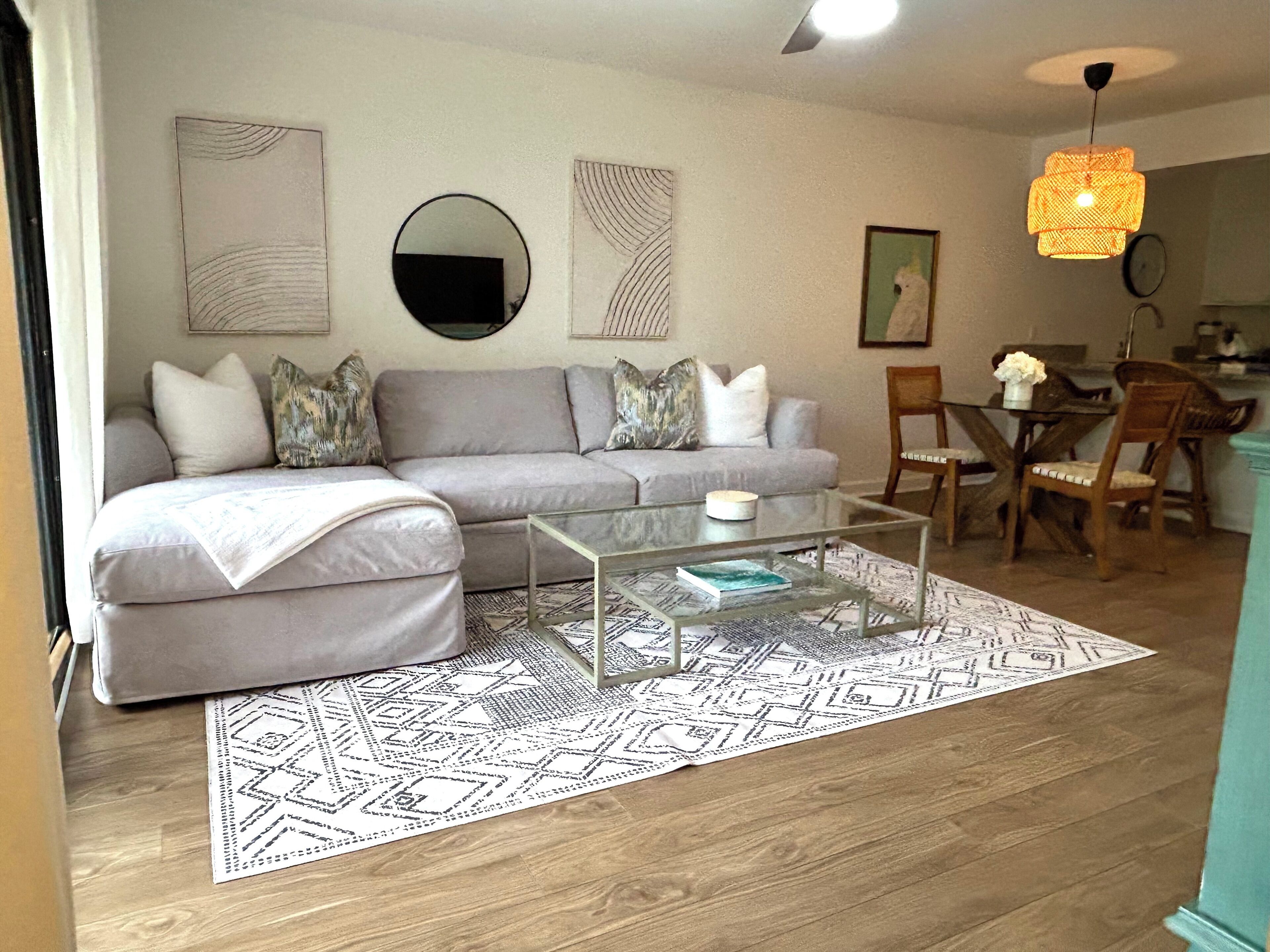 A living room with a sofa, coffee table, and patterned rug. There are wall art pieces and a round mirror. A dining area with chairs and a table is visible in the background.