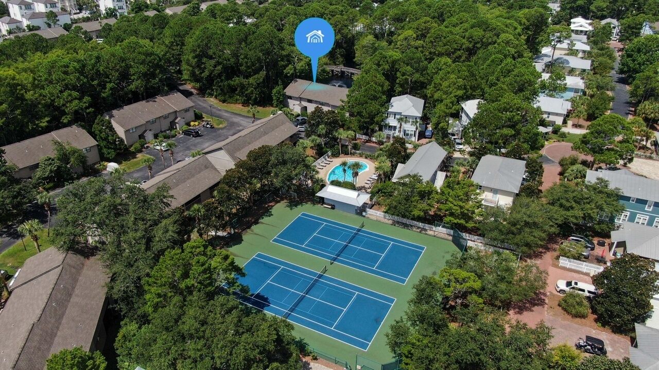 Vue aérienne d’un quartier résidentiel avec un court de tennis entouré de maisons et de verdure.
