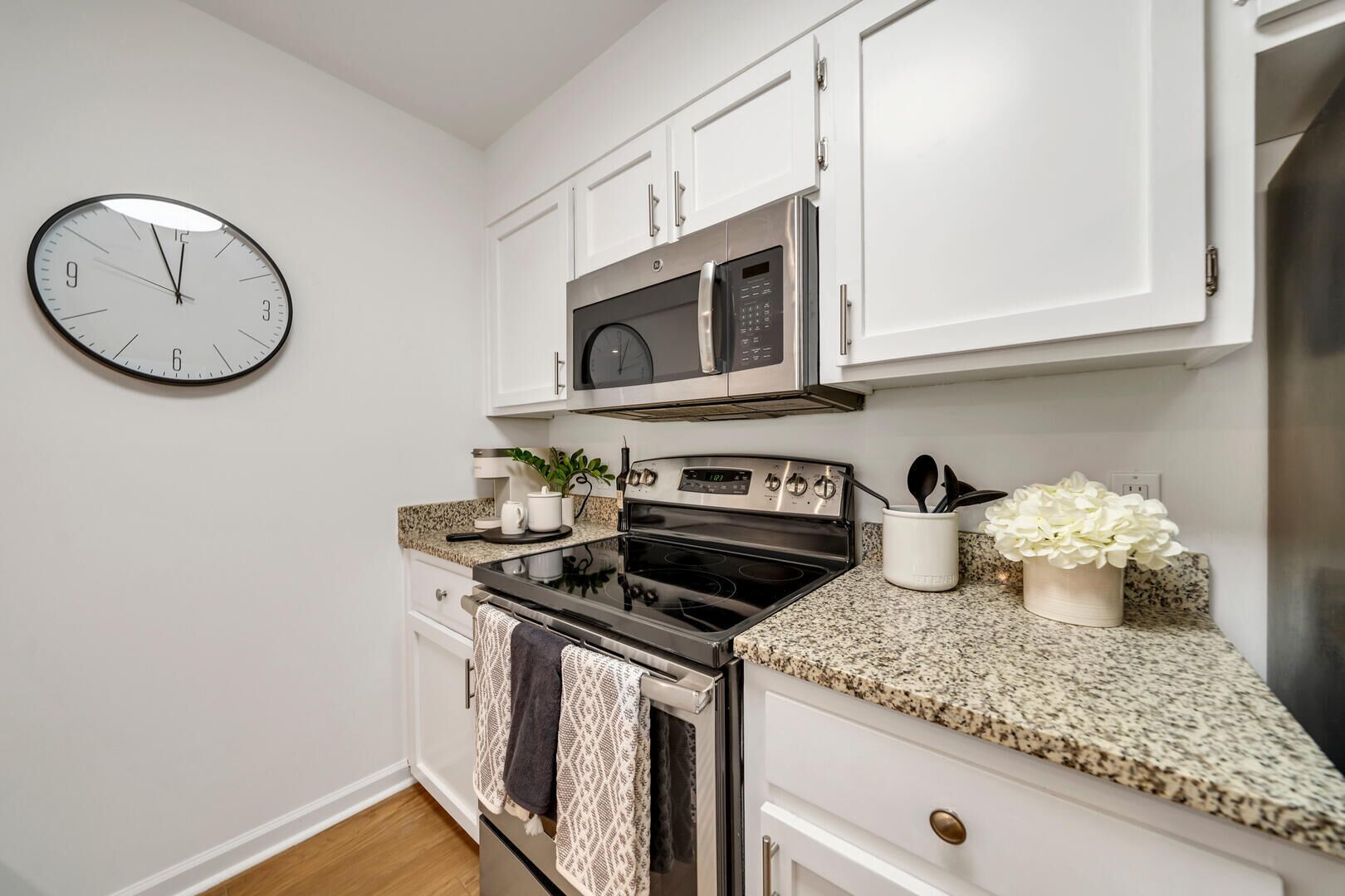 A kitchen with white cabinets, a stainless steel microwave, an oven, and a clock on the wall.