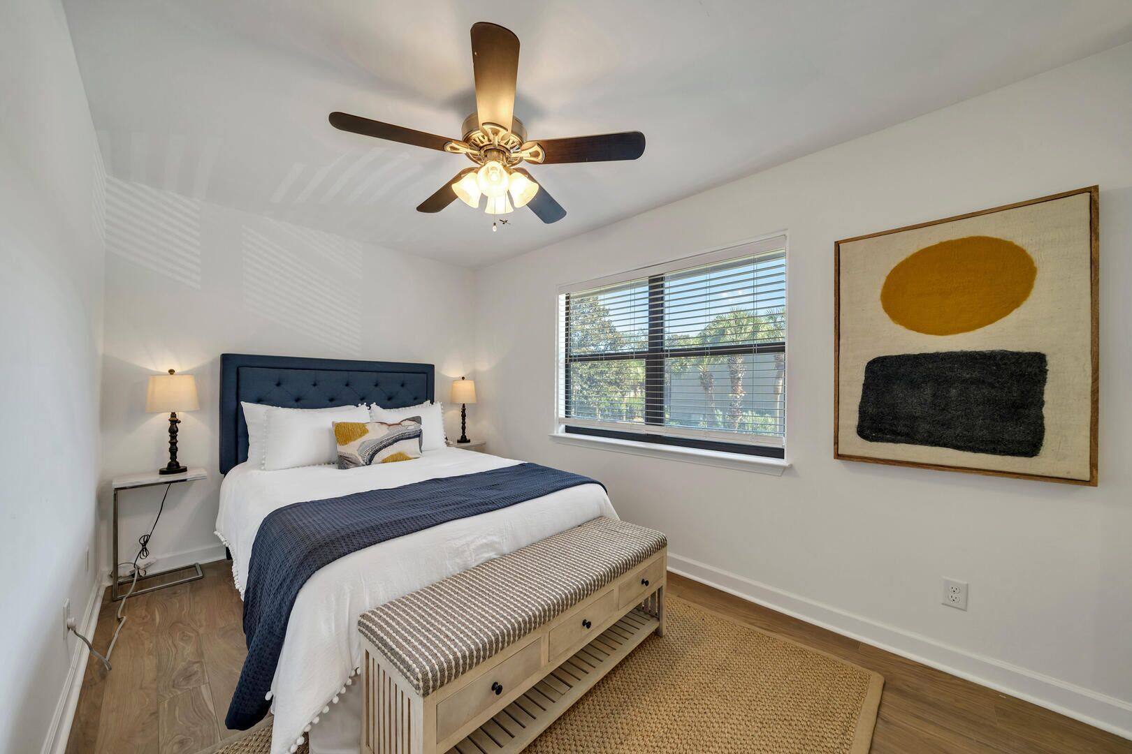 A bedroom with a bed, bedside lamps, a ceiling fan, a window with blinds, and a painting on the wall.