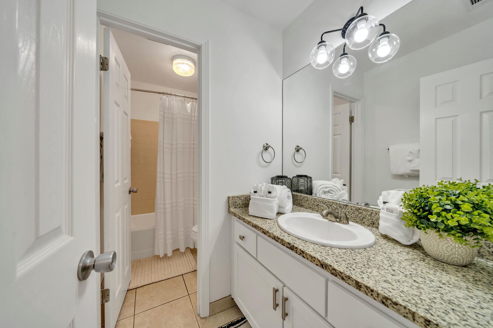 A bathroom with a white sink, granite countertop, and a mirror. There is a shower curtain, a towel rack, and a potted plant on the counter.