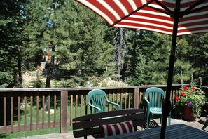 Relax on the patio surrounded by lush trees, with a comfortable bench and two chairs under a shaded umbrella.