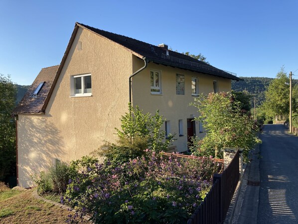 A two-story house with a sloped roof, a small porch, and a garden with purple flowers.